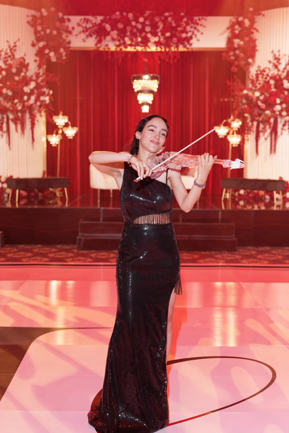 Sadie Varius in a black sequined gown, the transparent violin on a red-lit ballroom stage at The Legacy Castle
