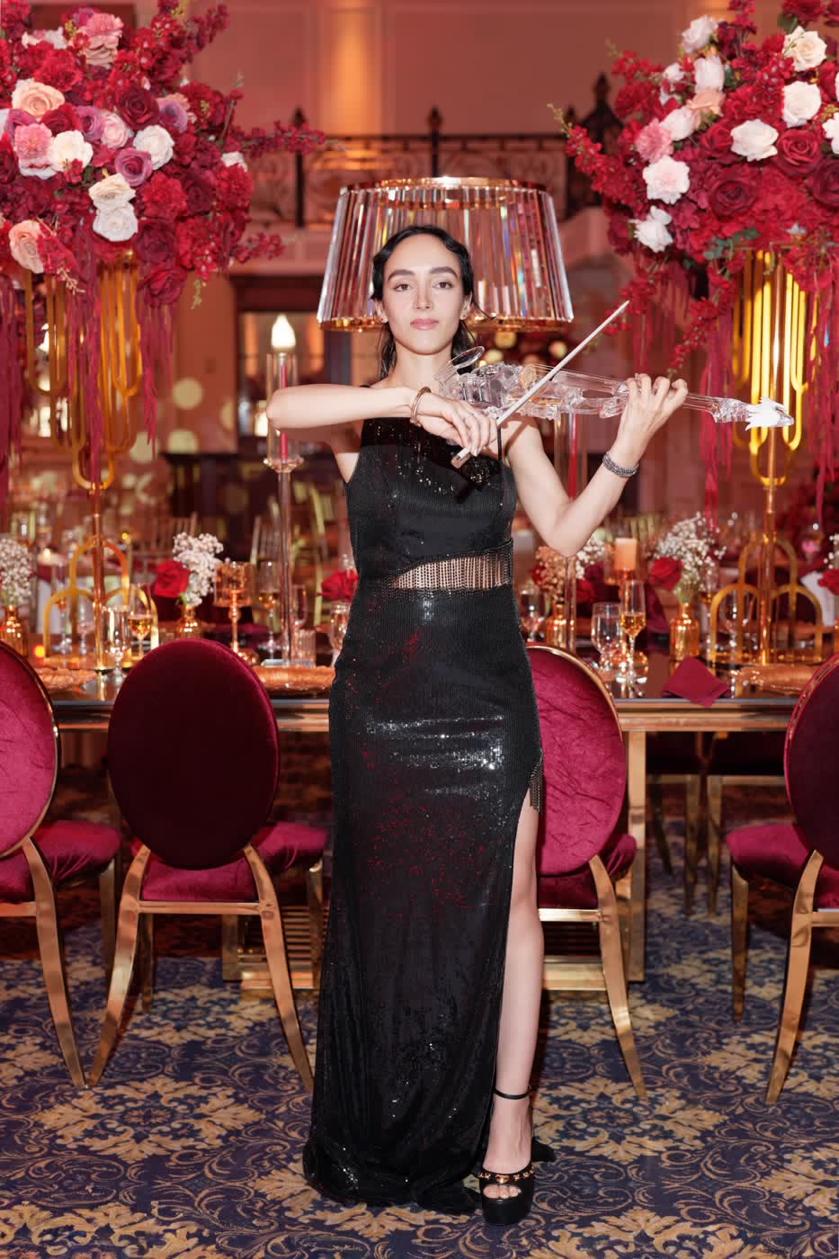 Sadie, bow raised, playing the transparent violin among towering rose centerpieces